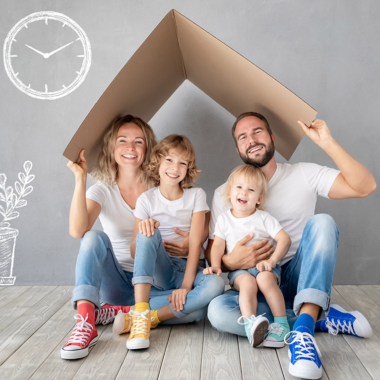 Eine Familie mit zwei Kindern sitzt fröhlich auf Holzboden vor grauer Wand, hält ein Pappdach über den Köpfen, trägt bunte Sneaker und weiße Shirts, Symbol für Geborgenheit und Zuhause.