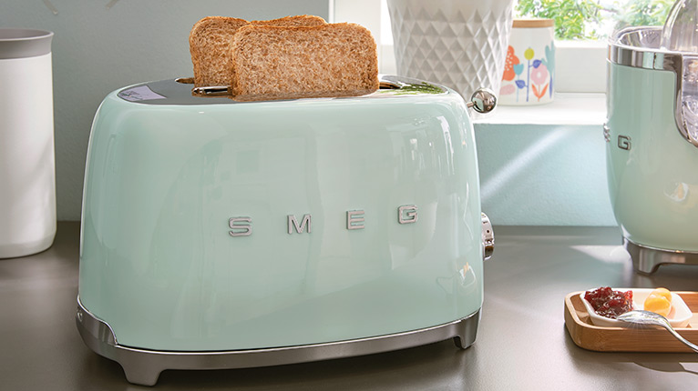 smeg Toaster bei Moebel-Shop – Mintgrüner Toaster mit 2 Toasts und ein Standmixer auf holzfarbener Arbeitsplatte vor einem Fenster, daneben steht ein Kuchenteller – farblich perfekt abgestimmt.