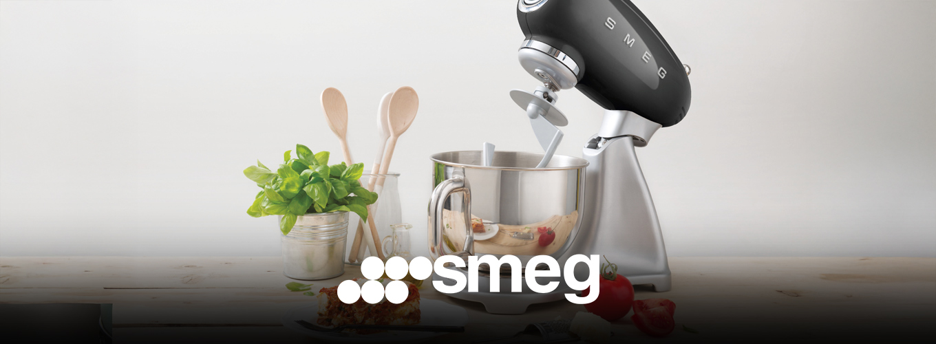 smeg Küchenmaschinen bei Moebel-Shop – Schwarze Küchenmaschine auf heller Holzarbeitsplatte. Umgeben von Kochutensilien, frischen Zutaten und einem angerichteten Teller Lasagne – Genuss trifft Stil.