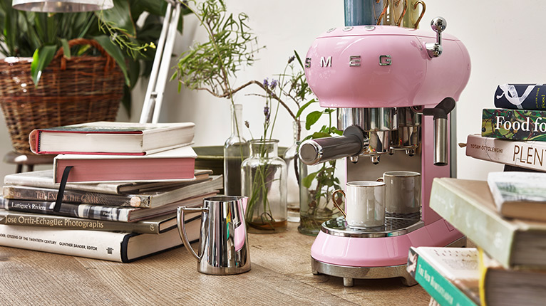 smeg Kaffeemaschinen bei Moebel-Shop – Rosa SMEG Espressomaschine mit Edelstahl-Milchkännchen daneben auf einer Arbeitsplatte mit Bücherstapeln daneben