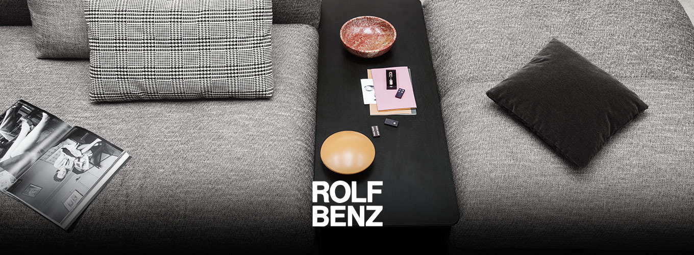 ROLF BENZ Accessoires - Zwei durch ein passendes, schwarzes Zwischentisch-Element unterbrochene graue ROLF BENZ Sofaelemente, auf denen Dekoration und Kissen zu sehen sind