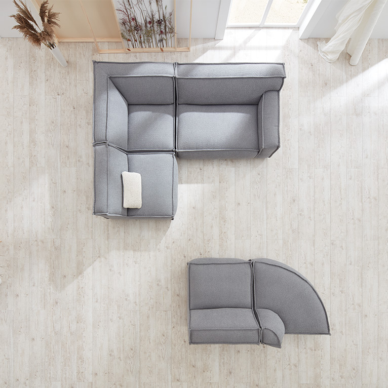 Modulares Sofa in L-Form und separatem Rundelement in hellgrauem Stoff, von oben fotografiert, auf hellem Holzfußboden in einem lichtdurchfluteten Wohnraum mit Dekoakzenten.
