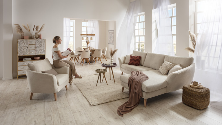 SANSIBAR LIVING Sofas bei Moebel-Shop - Ein in Creme und Beige gehaltenes Wohn-Esszimmer mit einem SANSIBAR Sofa und einem dazu passenden SANSIBAR Sessel, im Hintergrund  eine SANSIBAR Essgruppe und mehrere Sprossenfenster mit wehenden Gardinen
