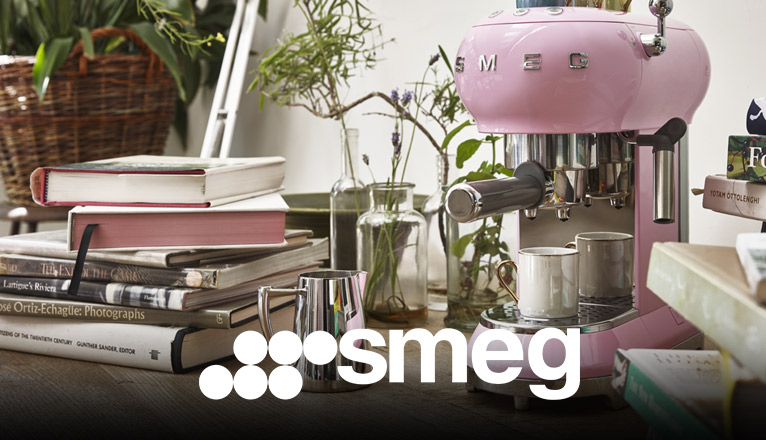 smeg Kaffeemaschinen bei Moebel-Shop – Rosa SMEG Espressomaschine mit Edelstahl-Milchkännchen daneben auf einer Arbeitsplatte mit Bücherstapeln daneben