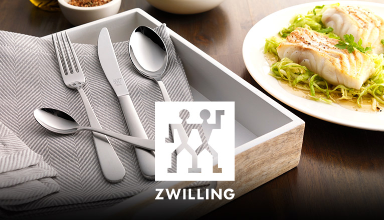 Großes Holztablett auf einem Tisch mit Stoffserviette in Taupe, einem Löffel, einem Messer, einer Gabel und einem Dessertlöffel eines ZWILLING-Bestecks; daneben ein Teller mit einem Lachs-Gericht.