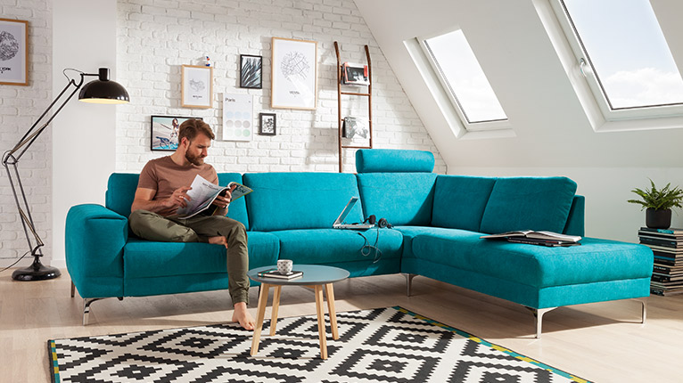 LIV’IN Polster bei Moebel-Shop - Ecksofa in Türkis mit verstellbarer Kopfstütze und filigranen Metallfüßen. Runder Beistelltisch aus Holz und Teppich im grafischen Schwarz-Weiß-Muster ergänzen das moderne Ambiente.