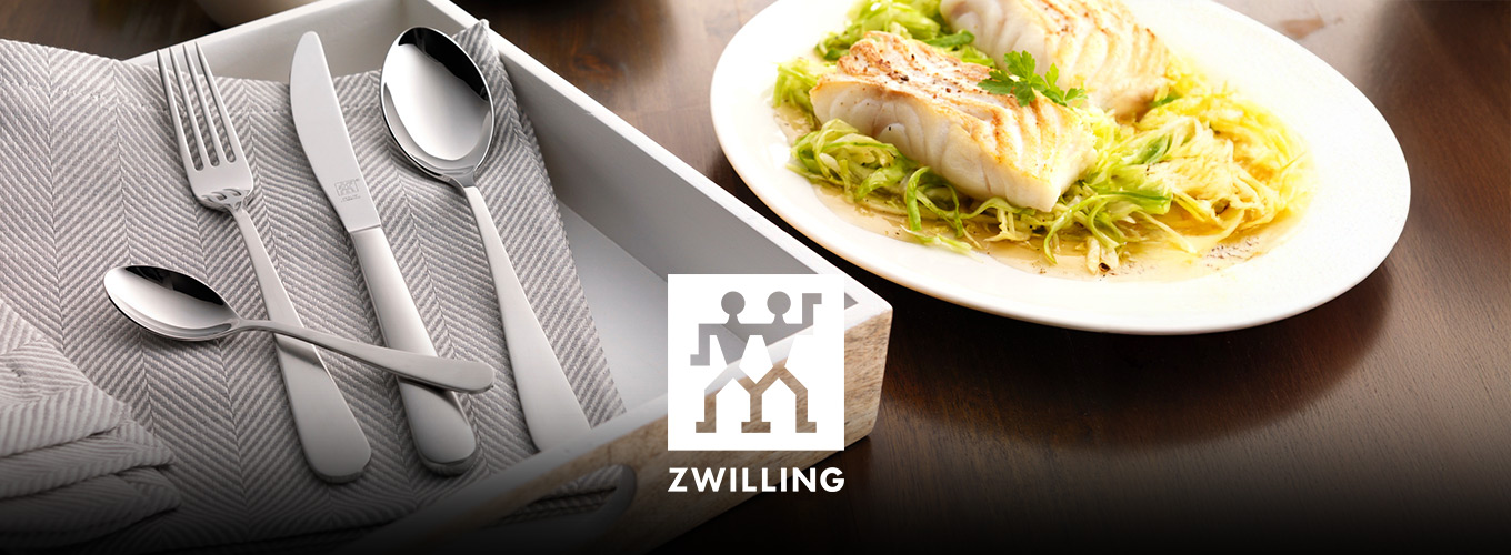 Großes Holztablett auf einem Tisch mit Stoffserviette in Taupe, einem Löffel, einem Messer, einer Gabel und einem Dessertlöffel eines ZWILLING-Bestecks; daneben ein Teller mit einem Lachs-Gericht.