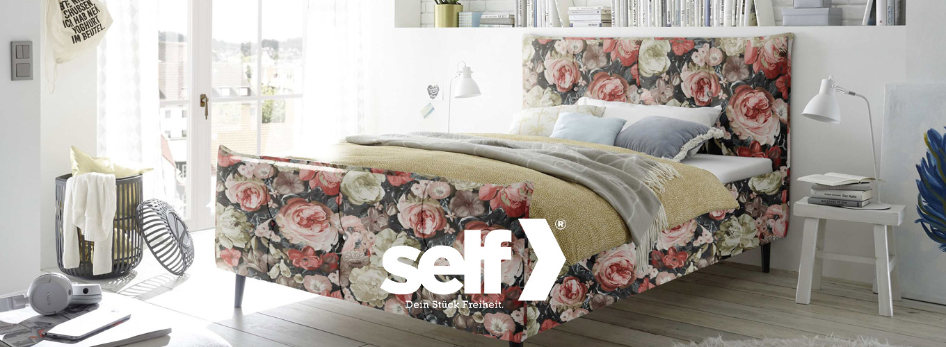self bei Moebel-Shop - Polsterbett mit floral gemustertem Stoffbezug an Kopf- und Fußteil. Das Bett steht in einem hellen Schlafzimmer mit modernen Beistelltischen, Bücherregal und Dekoration im Boho-Stil.