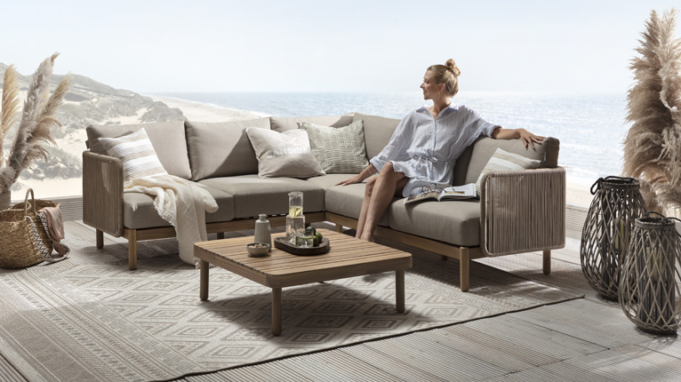 SANSIBAR LIVING Garten bei Moebel-Shop - Eine Frau in sommerlichem Blusenkleid sitzt auf einer SANSIBAR Garten-Eck-Lounge aus Holz und auf einer Terrasse mit einem plakativen Hintergrund, der eine Dünenlandschaft und das Meer zeigt