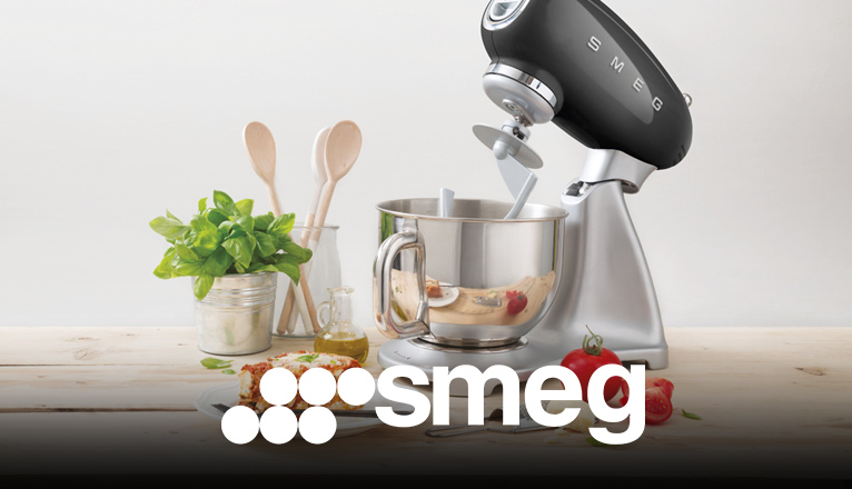 smeg Küchenmaschinen bei Moebel-Shop – Schwarze SMEG Küchenmaschine auf heller Holzarbeitsplatte. Umgeben von Kochutensilien, frischen Zutaten und einem angerichteten Teller Lasagne – Genuss trifft Stil.