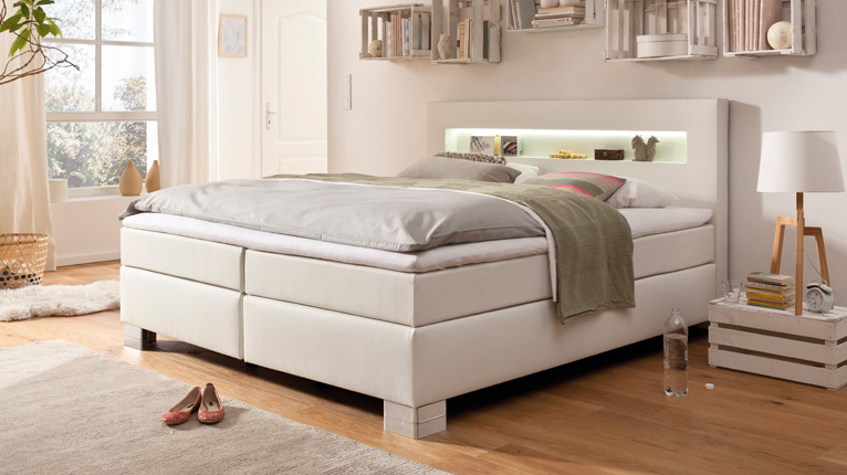 LIV’IN Schlafen bei Moebel-Shop - Weißes Boxspringbett mit beleuchtetem Kopfteil. Rustikale Wandregale aus Holzkisten ergänzen das gemütlich eingerichtete Schlafzimmer mit Holzfußboden.