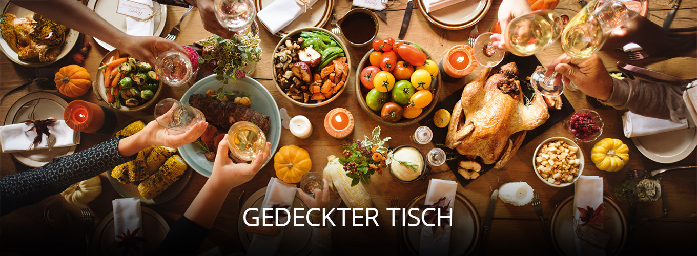 Festlich gedeckter Tisch mit vielfältigen Speisen wie Truthahn, Gemüse, Brot und Obst, dekoriert mit Kerzen und kleinen Kürbissen, während mehrere Personen gemeinsam anstoßen und feiern