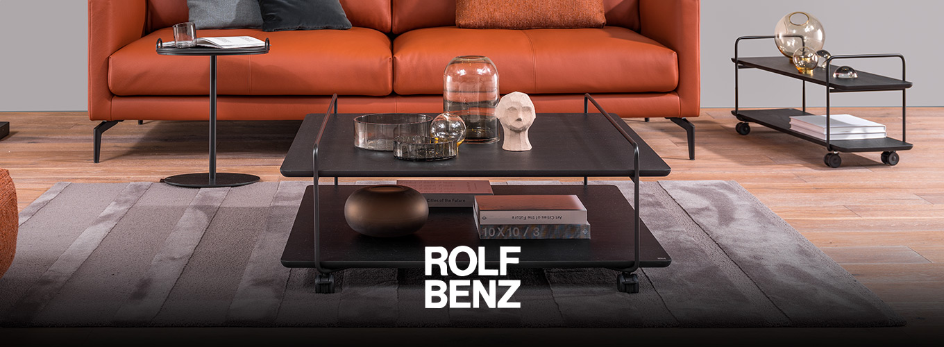 ROLF BENZ Couchtische - Ein schwarzer ROLF BENZ Couchtisch auf Rollen aus schwarzem Holz und Metall steht auf einem hochwertigen grau-silbernen Teppich vor einem prominenten ROLF BENZ Ledersofa in Orange