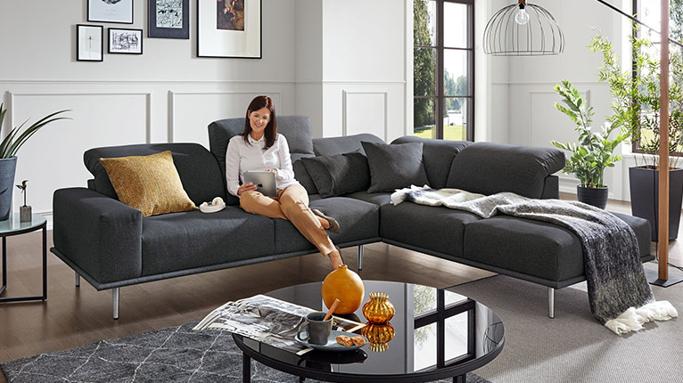 MONDO Sofas bei Moebel-Shop - Eine Frau sitzt entspannt auf einer großen, anthrazitfarbenen MONDO Polsterecke mit Metallfüßen und bedient ein iPad, in einem schönen Wohnzimmer mit gläsernem, schwarzen Couchtisch, Blumen und Bilder als Dekoration