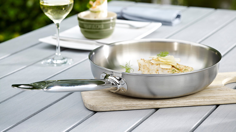 ZWILLING-Edelstahlpfanne mit gekochtem Essen auf einem Holzbrett auf dem Gartentisch; im Hintergrund gedeckter Platz mit Weinglas.