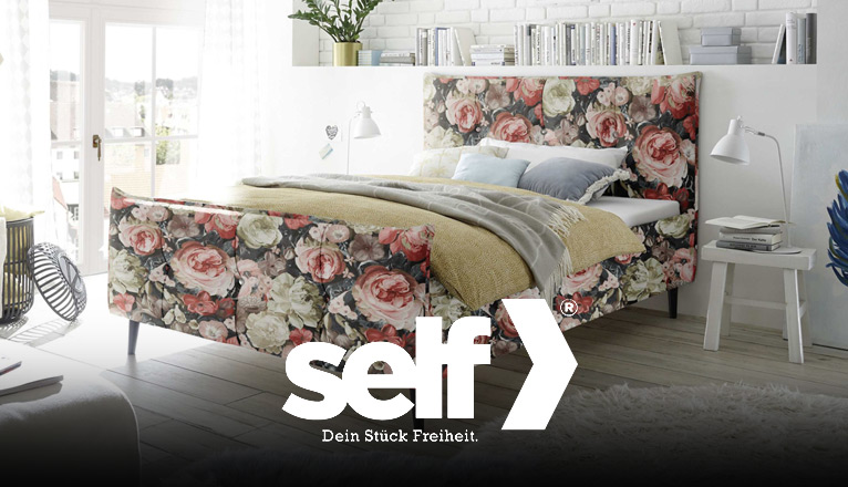 self bei Moebel-Shop - Polsterbett mit floral gemustertem Stoffbezug an Kopf- und Fußteil. Das Bett steht in einem hellen Schlafzimmer mit modernen Beistelltischen, Bücherregal und Dekoration im Boho-Stil.