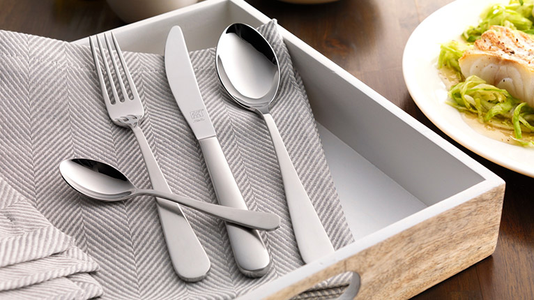 Großes Holztablett auf einem Tisch mit Stoffserviette in Taupe, einem Löffel, einem Messer, einer Gabel und einem Dessertlöffel eines ZWILLING-Bestecks; daneben ein Teller mit einem Lachs-Gericht.