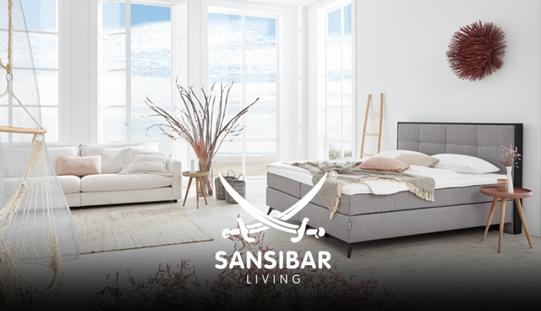 SANSIBAR LIVING Schlafen bei Moebel-Shop - Ein silbernes SANSIBAR Boxspringbett in einem hellen Schlafzimmer mit bodentiefen Sprossenfenstern und einem cremefarbenen SANSIBAR Sofa und natürlicher Dekoration und Blumen in zartem Rosa