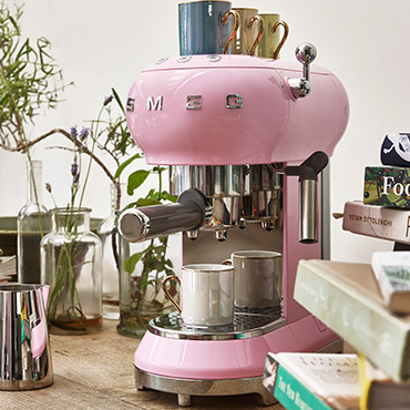 smeg Kaffeemaschinen bei Moebel-Shop – Rosa SMEG Espressomaschine mit Edelstahl-Milchkännchen daneben auf einer Arbeitsplatte mit Bücherstapeln daneben