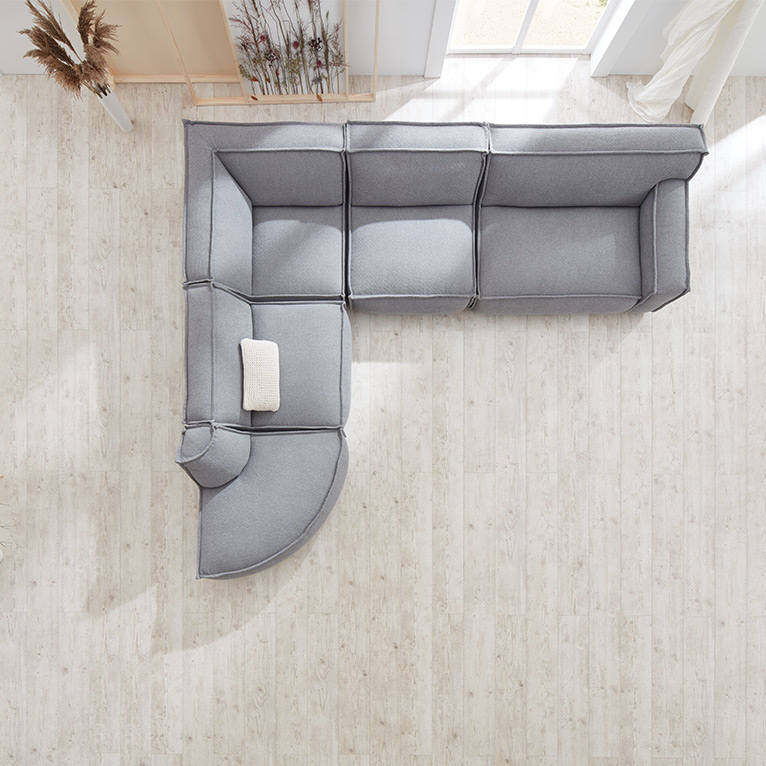 L-förmiges, hellgraues Sofa mit runder Ecke und heller Zierkissenauflage, von oben aufgenommen auf hellem Holzboden in einem hell eingerichteten Raum mit viel Tageslicht.