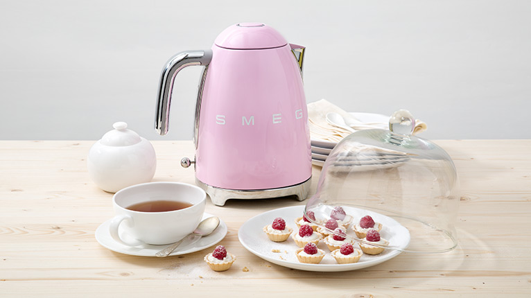 smeg Wasserkocher bei Moebel-Shop – Rosa SMEG Wasserkocher mit Teetasse, Zuckerdose und passenden, weißen Tellern. Auf dem Teller im Vordergrund liegen kleine Himbeertörtchen eine Glasabdeckung liegt halb darauf