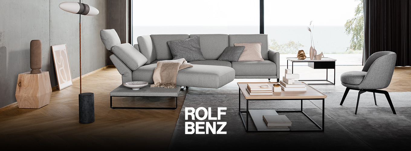 ROLF BENZ Sofas - Ein hellgraues ROLF BENZ Sofa mit schwenkbarem Liegeelement, ein ROLF BENZ Sessel und ROLF BENZ Couchtische in einem Wohnzimmer mit großer Fensterfront und Wänden in Beton-Optik