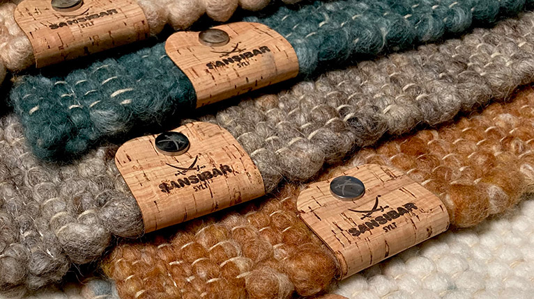 SANSIBAR LIVING Teppiche bei Moebel-Shop - Mehrere SANSIBAR Teppiche mit SANSIBAR Label in Petrol, Braun, Creme und Schlamm liegen im Bildanschnitt übereinander