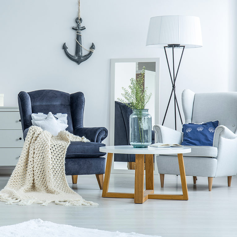Wohnzimmer im maritimen Stil mit Sesseln, hellem Couchtisch, Stehlampe, Anker-Wanddekoration und Wohnaccessoires in Blau- und Naturtönen.