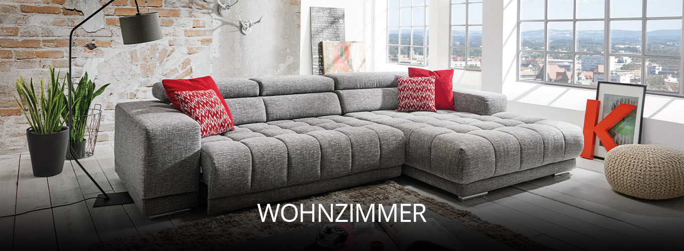 Helles Wohnzimmer mit großer grauer Eckcouch, roten Kissen als Farbakzent, dekorativen Elementen und Pflanzen, kombiniert mit loftartiger Backsteinwand und Panoramafenstern für urbanes Flair
