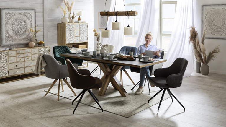 SANSIBAR LIVING Speisen bei Moebel-Shop - Eine SANSIBAR Essgruppe mit SANSIBAR Esstisch und unterschiedlich farbigen SANSIBAR Polsterstühlen, eine Frau sitzt am Tisch und blättert in einem Katalog, natürliche Dekoration und SANSIBAR Möbel im Hintergrund