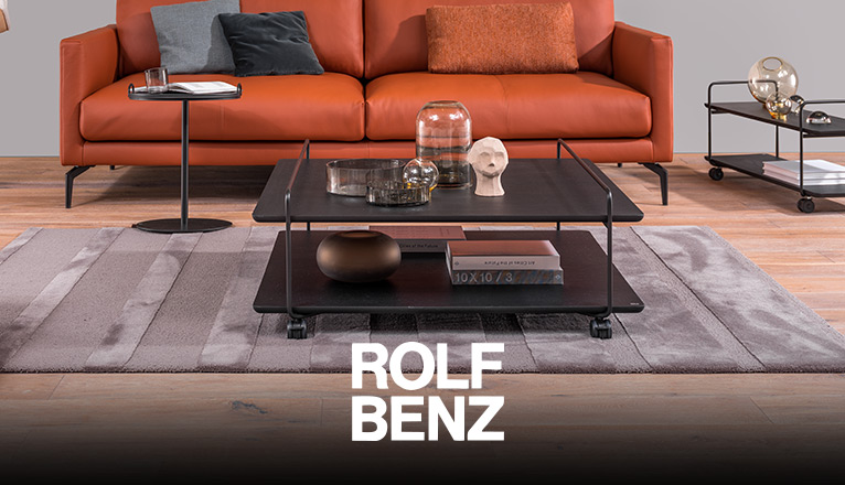 ROLF BENZ Couchtische - Ein schwarzer ROLF BENZ Couchtisch auf Rollen aus schwarzem Holz und Metall steht auf einem hochwertigen grau-silbernen Teppich vor einem prominenten ROLF BENZ Ledersofa in Orange