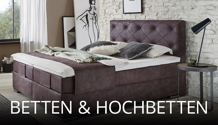 Komfortables Schlafzimmer mit großem Polsterbett in dunklem Stoff, dekorativen Kissen und Plaid, kombiniert mit Steinwand, seitlichem Beistelltisch und stimmungsvollem Tageslicht für gemütliches Ambiente