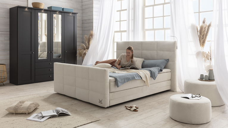 SANSIBAR LIVING Schlafen bei Moebel-Shop - Ein cremefarbenes SANSIBAR Boxspringbett in einem hellen Schlafzimmer mit bodentiefen Sprossenfenstern und einem graphit-schwarzem Kleiderschrank, zwei passende SANSIBAR Poufs stehen neben dem Bett