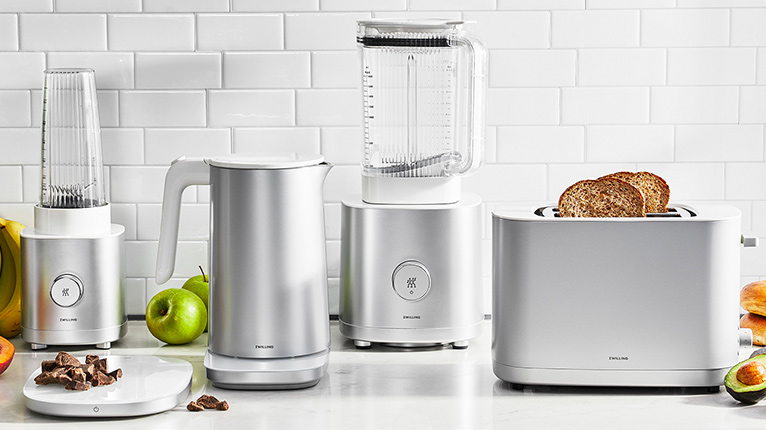 ZWILLING Enfinigy-Serie mit kleinem Standmixer, Wasserkocher, großem Standmixer und Toaster; umgeben von Obst und Brot.