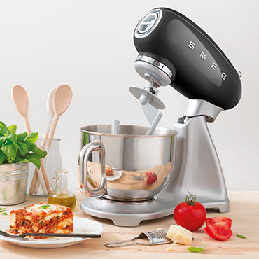 smeg Küchenmaschinen bei Moebel-Shop – Schwarze Küchenmaschine auf heller Holzarbeitsplatte. Umgeben von Kochutensilien, frischen Zutaten und einem angerichteten Teller Lasagne – Genuss trifft Stil.