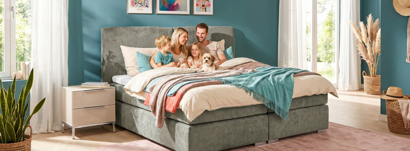 Familie entspannt gemeinsam auf einem Boxspringbett mit Hund in einem hellen Schlafzimmer mit modernen Möbeln und wohnlichen Dekorationen.