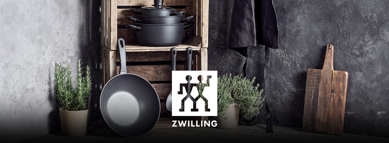 ZWILLING bei Moebel-Shop - ZWILLING-Kochgeschirr-Set mit schwarzen Töpfen und Pfannen vor rustikalem Holzregal, umgeben von Kräutertöpfen, Kochutensilien, Schneidebrett und Schürze.