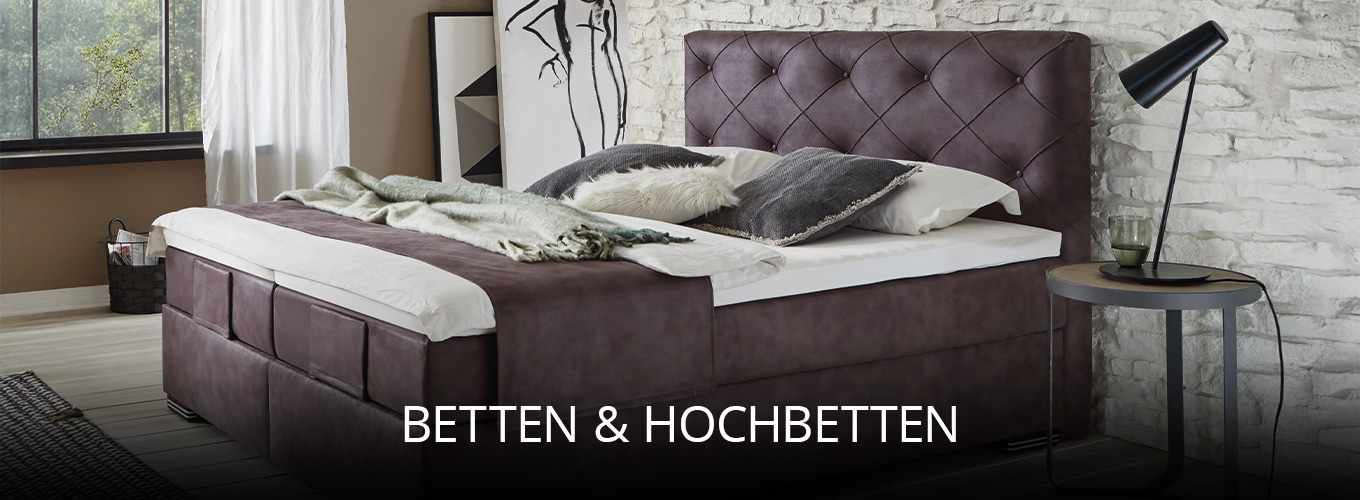 Komfortables Schlafzimmer mit großem Polsterbett in dunklem Stoff, dekorativen Kissen und Plaid, kombiniert mit Steinwand, seitlichem Beistelltisch und stimmungsvollem Tageslicht für gemütliches Ambiente