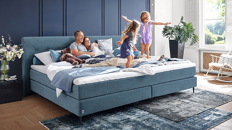 MONDO Schlafen bei Moebel-Shop - Eine Familie mit zwei Kindern in einem blauen MONDO Familien-Boxspringbett. Die Eltern liegen Arm in Arm und schauen lachend zu, wie ihre Kinder auf dem Bett freudestrahlend hüpfen.