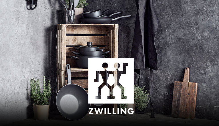 ZWILLING bei Moebel-Shop - ZWILLING-Kochgeschirr-Set mit schwarzen Töpfen und Pfannen vor rustikalem Holzregal, umgeben von Kräutertöpfen, Kochutensilien, Schneidebrett und Schürze.