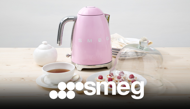 smeg Wasserkocher bei Moebel-Shop – Rosa SMEG Wasserkocher mit Teetasse, Zuckerdose und passenden, weißen Tellern. Auf dem Teller im Vordergrund liegen kleine Himbeertörtchen eine Glasabdeckung liegt halb darauf