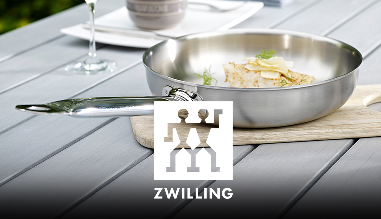 ZWILLING-Edelstahlpfanne mit gekochtem Essen auf einem Holzbrett auf dem Gartentisch; im Hintergrund gedeckter Platz mit Weinglas.