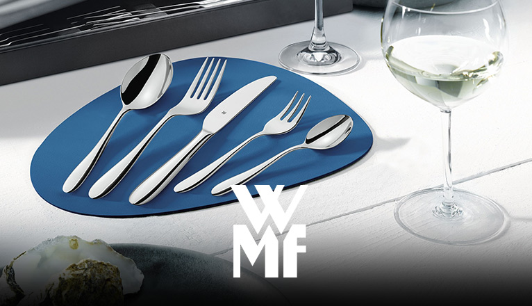 WMF-Besteckset aus fünf Teilen – Suppenlöffel, Gabel, Messer, Kuchengabel und Kaffeelöffel – auf blauer Tischunterlage; daneben steht ein Weinglas.