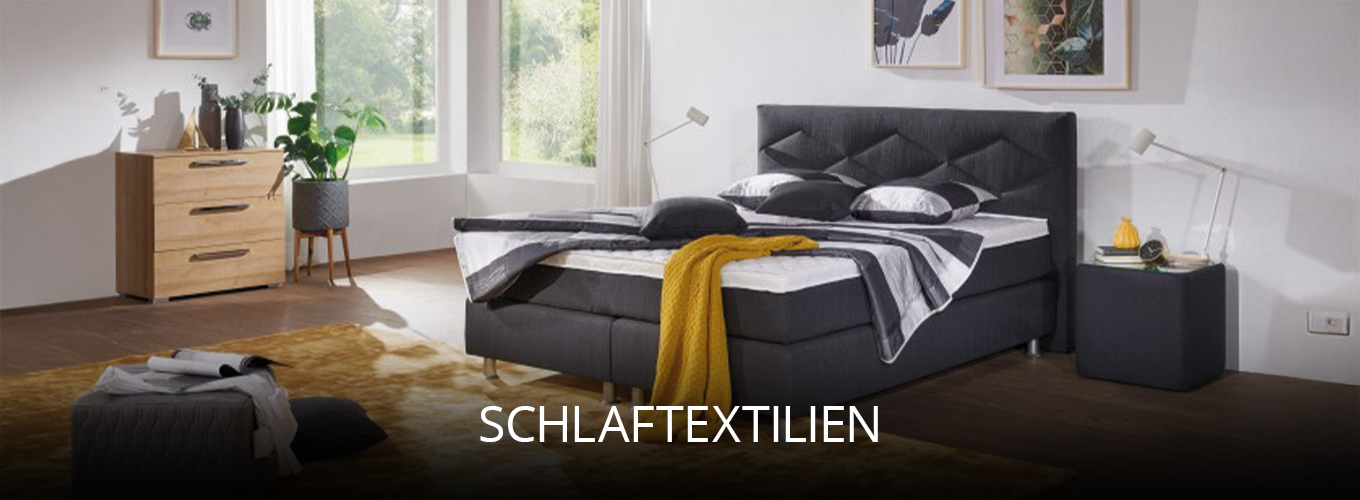 Gemütliches Schlafzimmer mit grauem Boxspringbett, dekorativen Kissen und Decken in kontrastreichen Farben, ergänzt durch Teppich, Kommode und große Fenster, die für viel Tageslicht sorgen