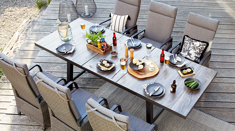 outdoor Gartentische bei Moebel-Shop - Ein von oben fotografierter outdoor Gartentisch aus Holz mit passenden outdoor Gartenstühlen mit grauen Polstern, Tischdekoration, Essen und Getränke auf dem Tisch