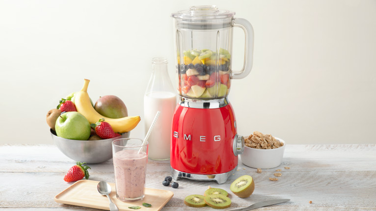 smeg Standmixer bei Moebel-Shop – Roter SMEG Standmixer mit einem gemixten Smoothie in einem Glas mit Strohhalm, eine Schale mit Obst, eine Flasche Milch und eine Schale mit Cerealien stehen daneben