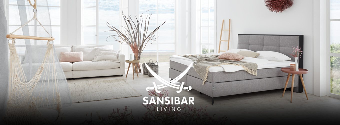 SANSIBAR LIVING Schlafen bei Moebel-Shop - Ein silbernes SANSIBAR Boxspringbett in einem hellen Schlafzimmer mit bodentiefen Sprossenfenstern und einem cremefarbenen SANSIBAR Sofa und natürlicher Dekoration und Blumen in zartem Rosa