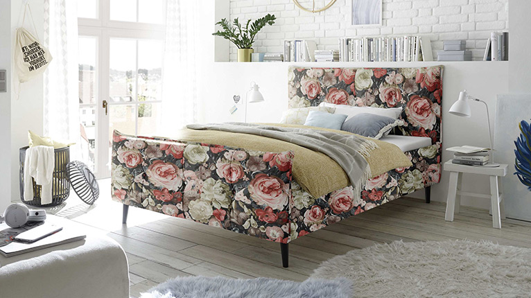 self Schlafen bei Moebel-Shop - Schlafzimmer mit floral gemustertem Polsterbett, weißen Kleiderschränken, Nachttisch im Retro-Stil und Wandregalen. Gemütliches Wohnkonzept mit dekorativer Farbgestaltung.