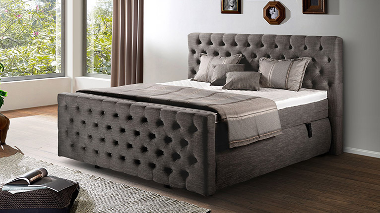 vito Schlafen bei Moebel-Shop - Ein glänzend taupefarbenes vito Boxspringbett in einem Schlafzimmer mit großen Fenstern und passenden Vorhängen