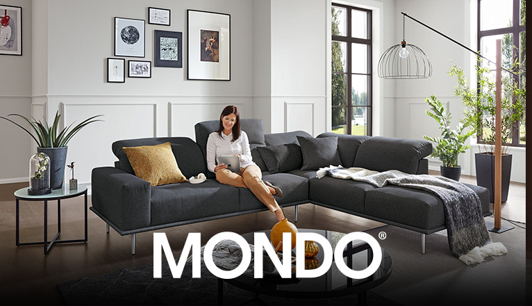 MONDO Sofas bei Moebel-Shop - Eine Frau sitzt entspannt auf einer großen, anthrazitfarbenen MONDO Polsterecke mit Metallfüßen und bedient ein iPad, in einem schönen Wohnzimmer mit gläsernem, schwarzen Couchtisch, Blumen und Bilder als Dekoration